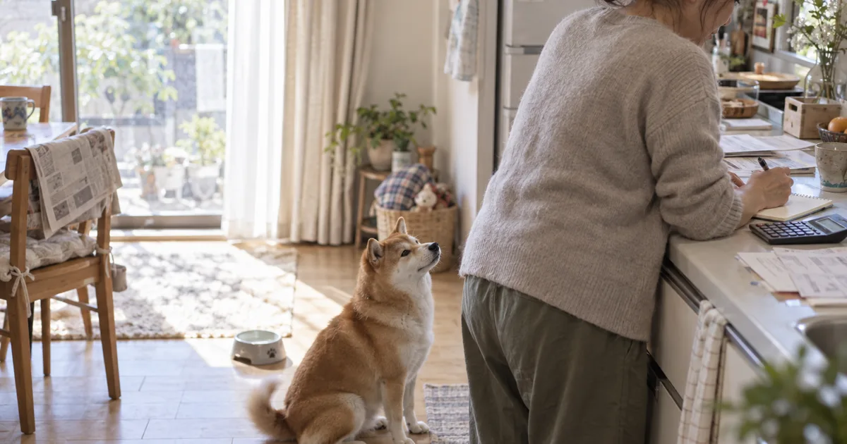 キッチンで費用をメモする50代の女性と、足元の柴犬